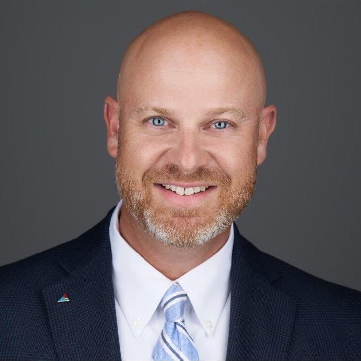 Mr. Kerry Bridges, a Caucasian male with a red and gray beard, smiles at the camera wearing a navy blue suit and is pictured against a grey background.