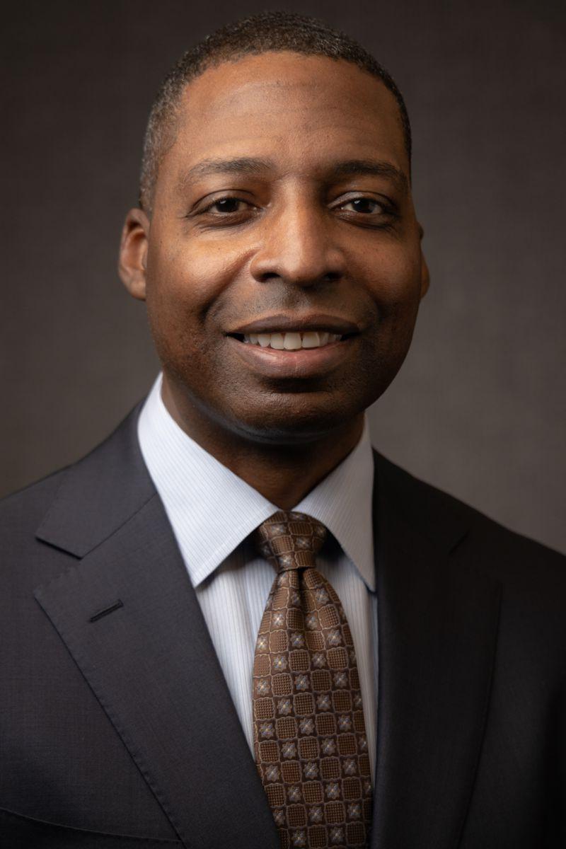 Mr. James Heffner, an African American male, wears a charcoal gray suit with a brown and tan patterned tie and is pictured against a gray background.