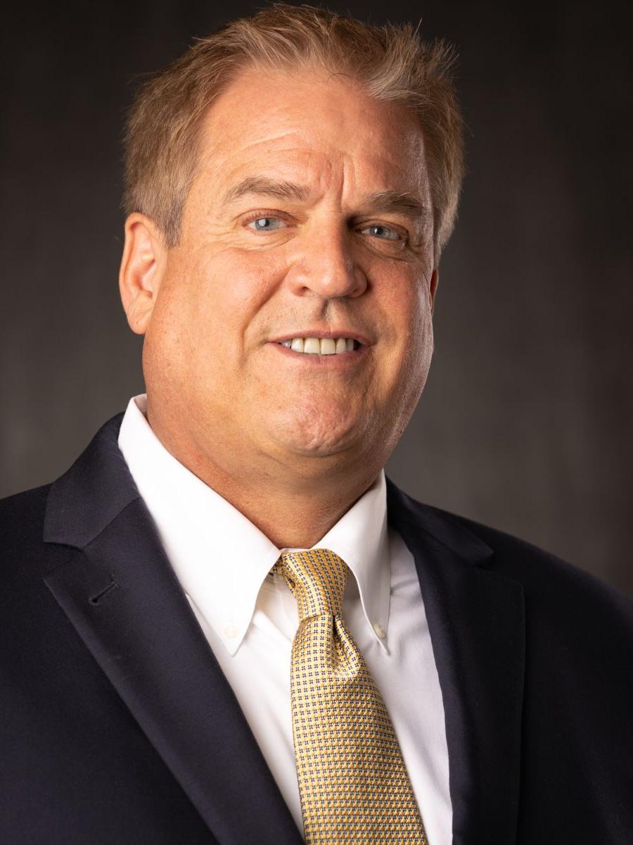 Mr. Jon Dollan, a Caucasian male with short grey and brown hair smile wearing a white collared shirt, gold tie with white dots, and a black suit jacket in front of a grey background.